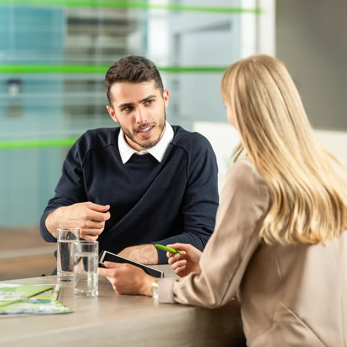 Foto einer Beratungssituation: Eine blonde langhaarige Frau berät einen Kunden - sie zeigt ihm Unterlagen auf einem Tablet.