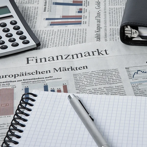 Foto von Zeitungen auf einem Tisch, darauf liegt ein Taschenrechner, ein karierter Block mit Stift, eine Aktentasche und ein Handy.