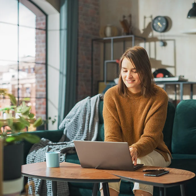 Eine junge Frau mit senf-farbenem Pullower und schulterlangen braunen Haaren auf einer Couch, am Tisch vor ihr steht ein Laptop.